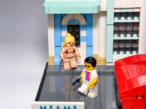 Zwei LEGO-Figuren vor einem Gebäude in Miami, eine Figur sitzt draußen und trägt einen Badeanzug, die andere Figur trägt eine weiße Jacke und hält eine Waffe.
